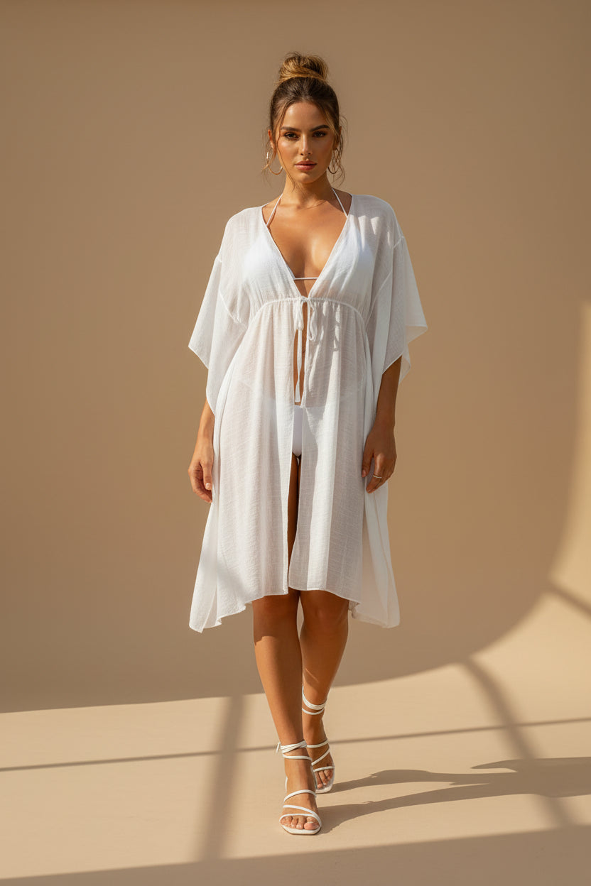 Woman in a white bikini and cover-up standing by a pool with a white building in the background.